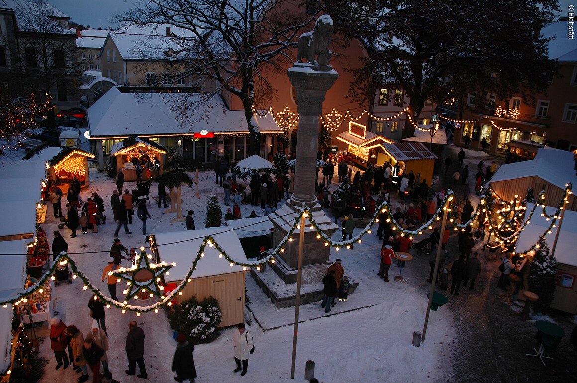Ein verschneiter Weihnachtsmarkt mit beleuchteten Ständen und einem zentralen Denkmal, umgeben von Menschen.