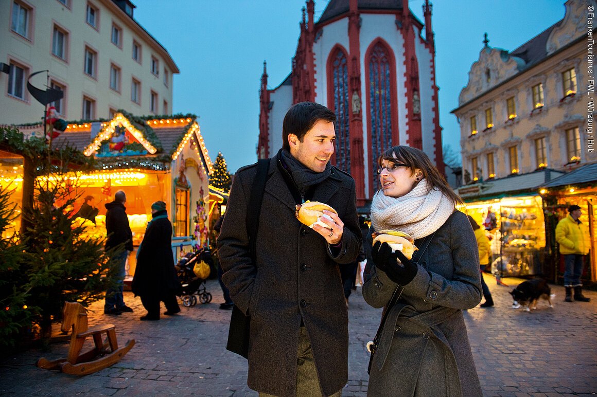 Zwei Personen essen Brötchen auf einem Weihnachtsmarkt mit beleuchteten Ständen und einer Kirche im Hintergrund.