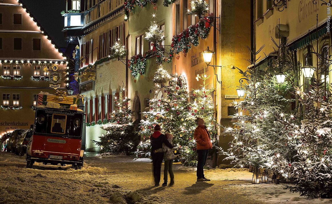 Verschneite Straße mit Weihnachtsbeleuchtung, geschmückten Tannenbäumen und Menschen, die spazieren gehen.