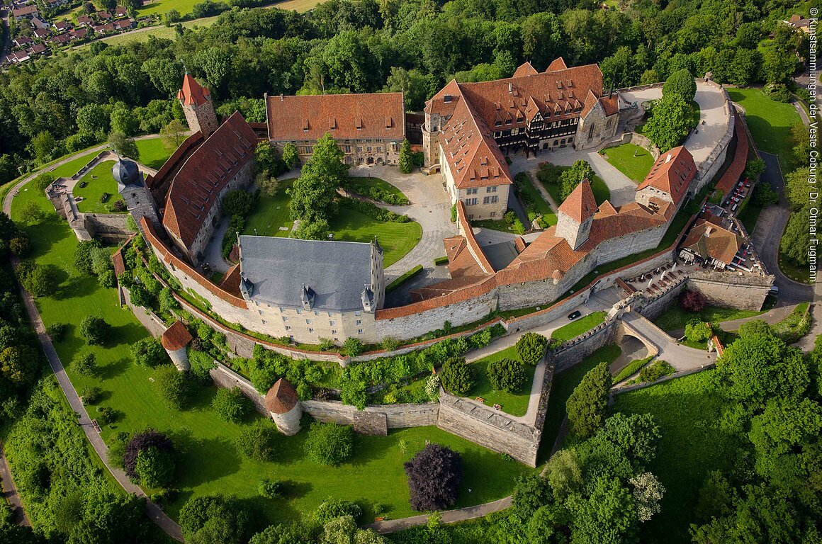 Luftaufnahme einer großen Burganlage mit roten Dächern, umgeben von grünen Bäumen und Rasenflächen.
