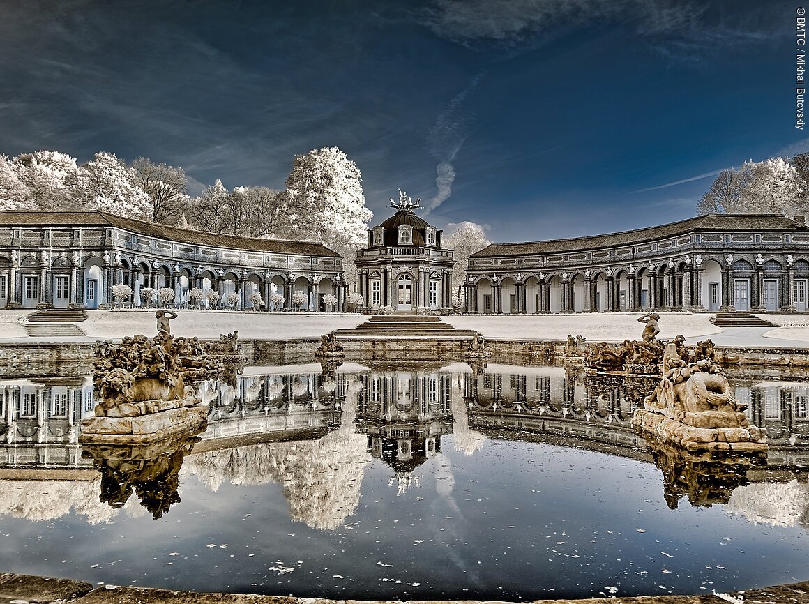 Historisches Gebäude mit Kuppel, flankiert von Kolonnaden, spiegelt sich in einem Teich wider. Skulpturen im Wasser.