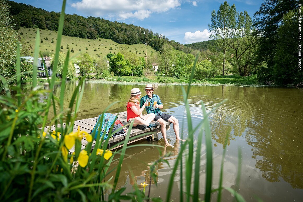 Zwei Personen sitzen auf einem Steg an einem See, umgeben von grüner Landschaft und gelben Blumen.
