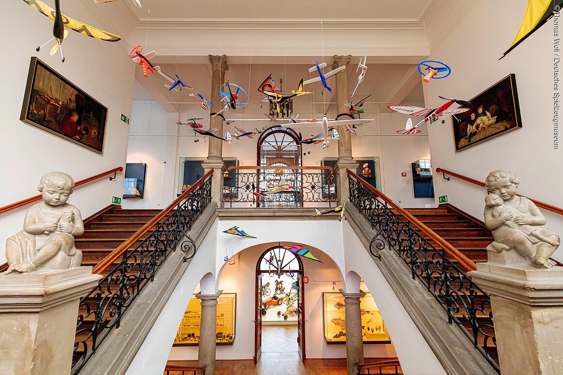 Innenansicht eines Museums mit Treppe, hängenden Modellflugzeugen und Skulpturen von Kindern.