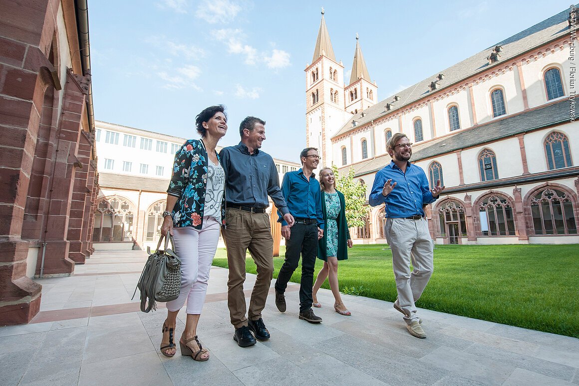 Fünf Personen gehen lächelnd durch einen Innenhof mit historischer Architektur und Rasenfläche.