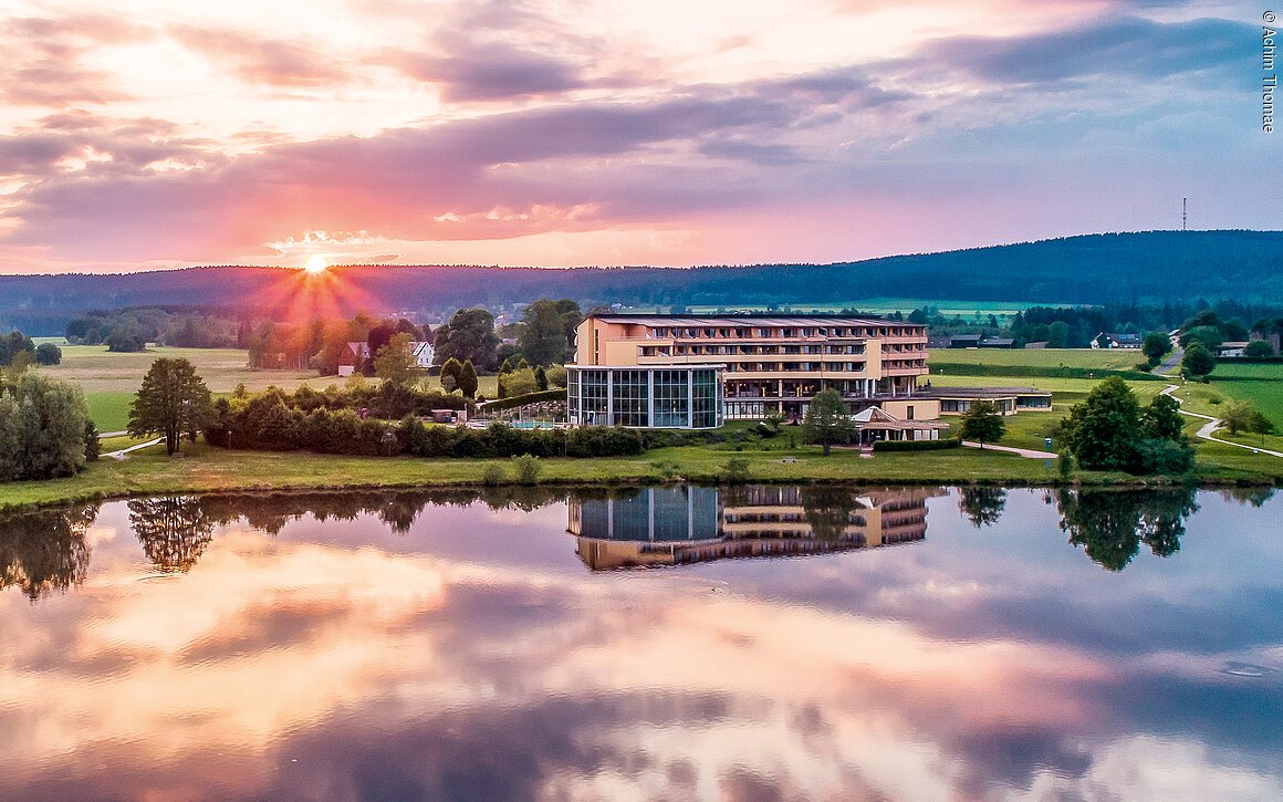Ein modernes Gebäude am Ufer eines Sees bei Sonnenuntergang, umgeben von grünen Feldern und Bäumen.