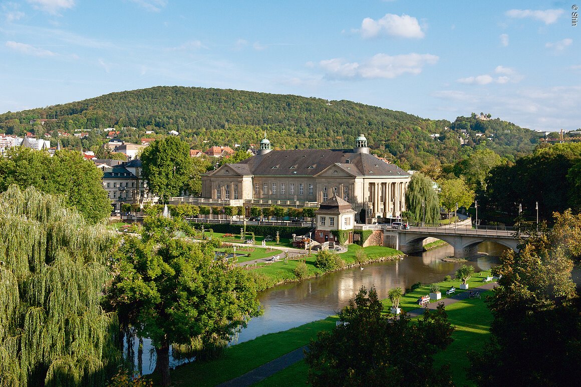 Gebäude mit Kuppeln und Säulen, umgeben von Bäumen und einem Fluss, im Hintergrund ein bewaldeter Hügel.