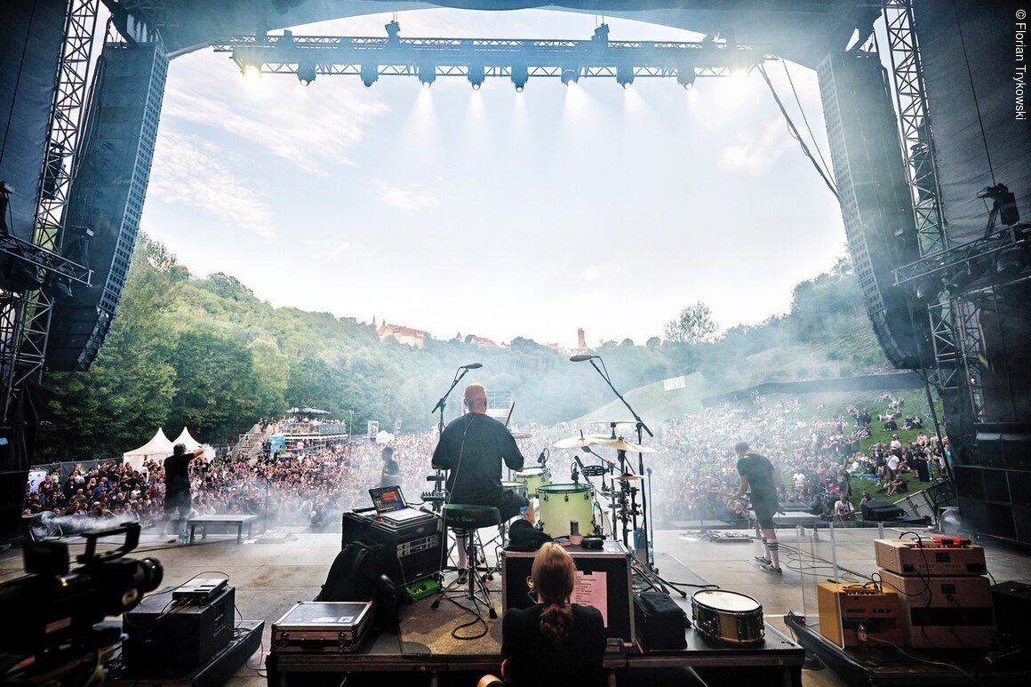 Blick von der Bühne auf Musiker und großes Publikum bei Open-Air-Konzert in grünem Tal am Tag.
